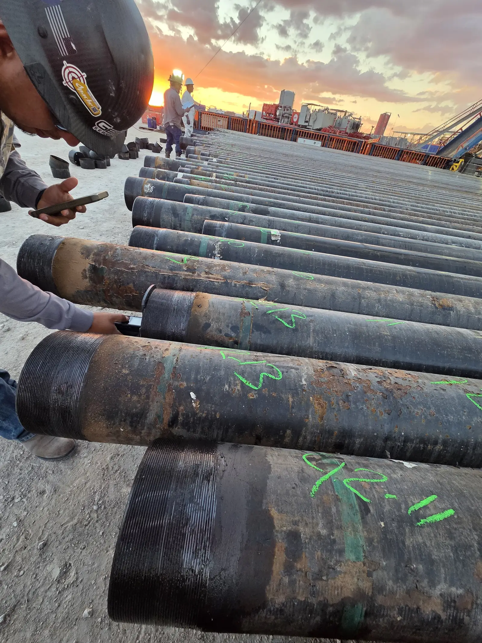 Gentex crew members in hard hats tallying casing on the pipe racks at sunset, two more of the crew and the rig behind them