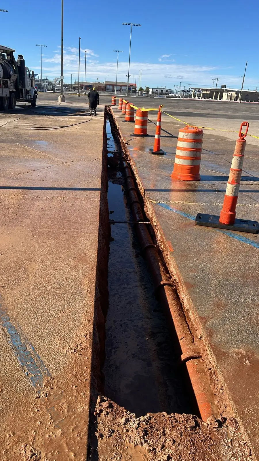 A hydro-excavated trench running across a paved parking lot, traffic barrels and caution tape set along it and the pavement edge cut clean