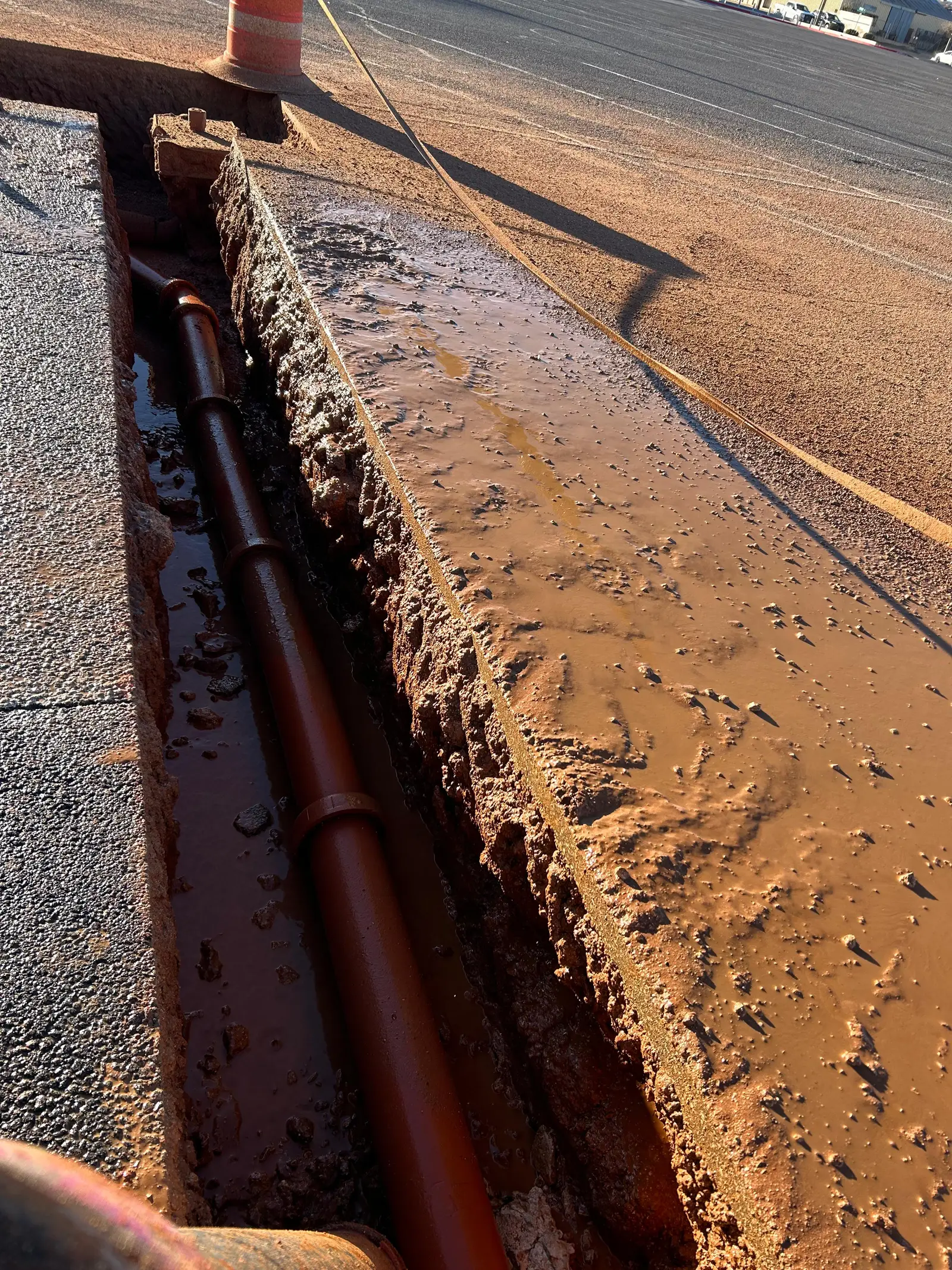 A long, straight hydro-excavated trench cut right along the edge of a paved lot, pipe daylighted intact in the bottom and the wall left clean
