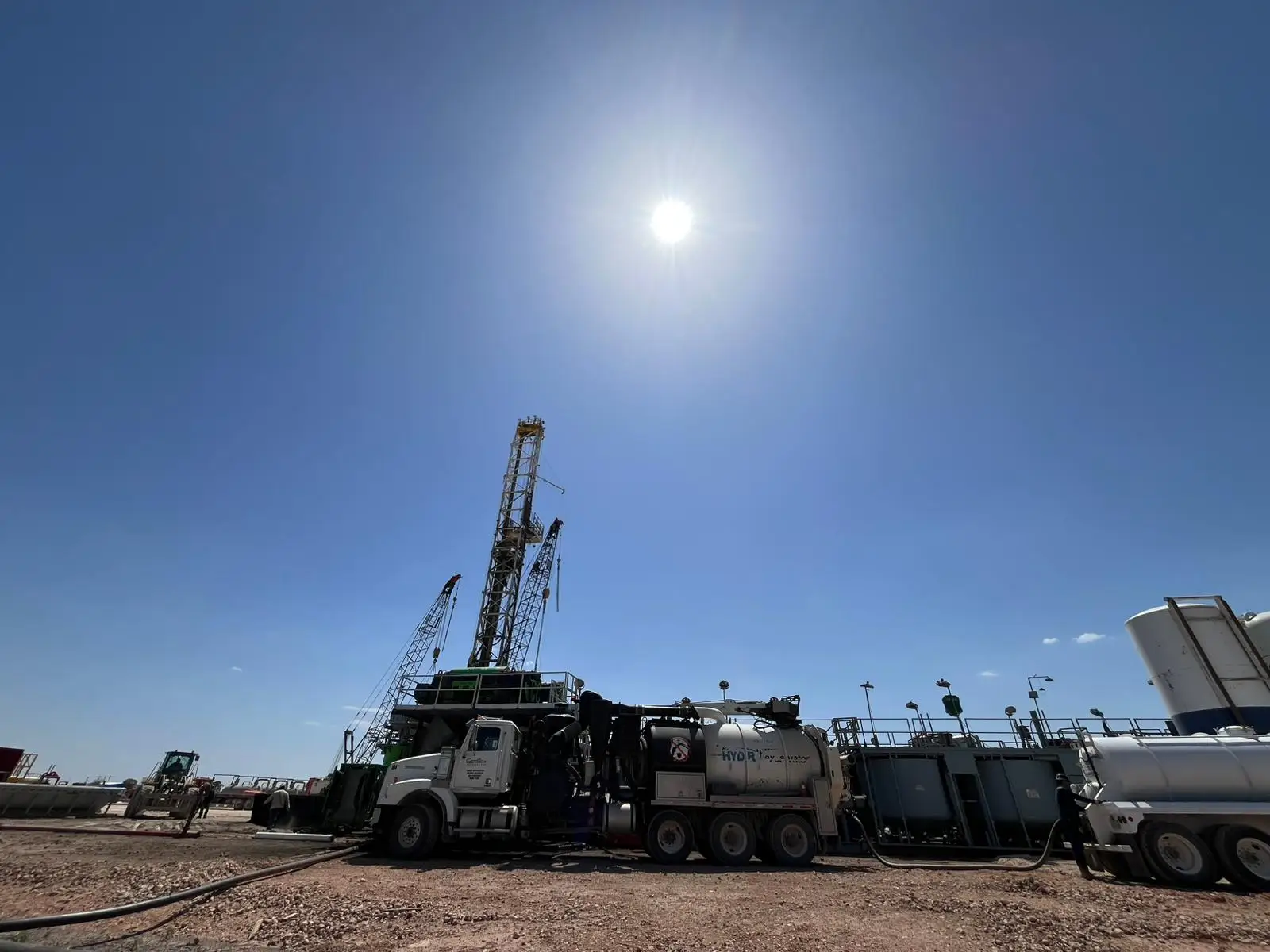 Gentex hydrovac unit rigged up beside a drilling rig on a Permian Basin location, suction line run out across the pad under a midday sun