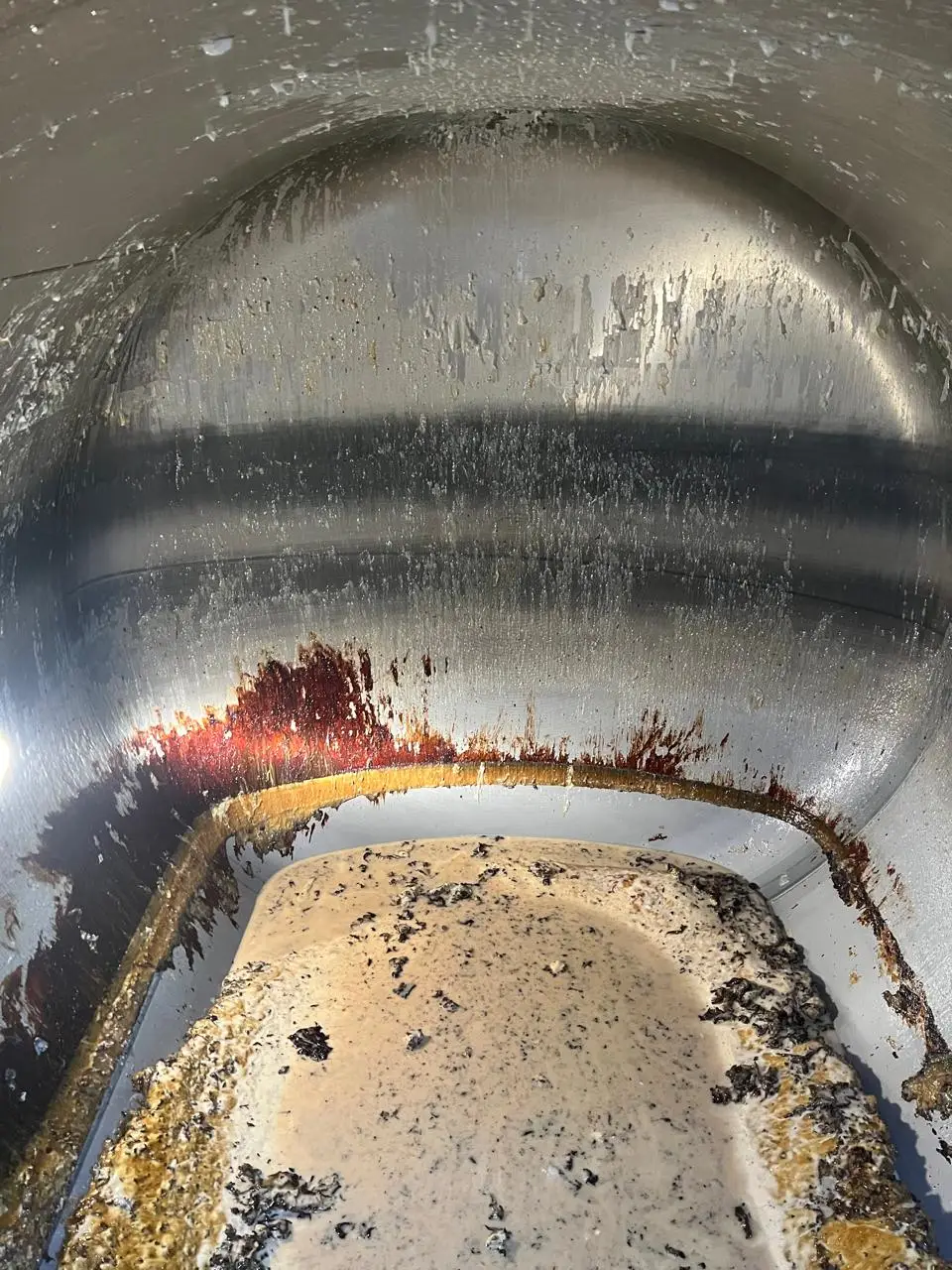 Inside an ISO tank before cleaning: rust-red and amber residue streaked down the head, with a crust of dried solids across the floor