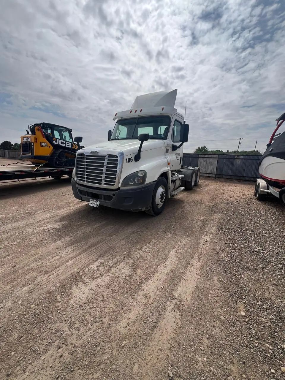A Gentex day cab tractor in the yard, staged to move a tanker or ISO container, with a skid steer loaded on a trailer behind it