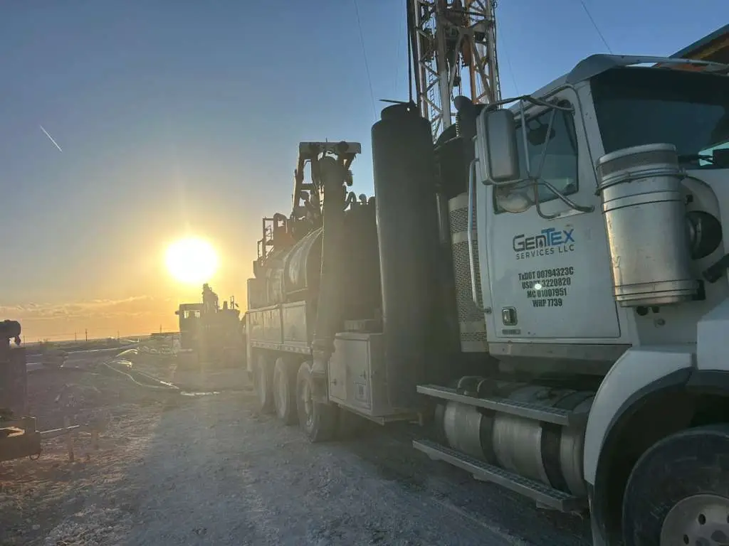 A Gentex vacuum truck working at first light, sun coming up behind a drilling rig on a West Texas location