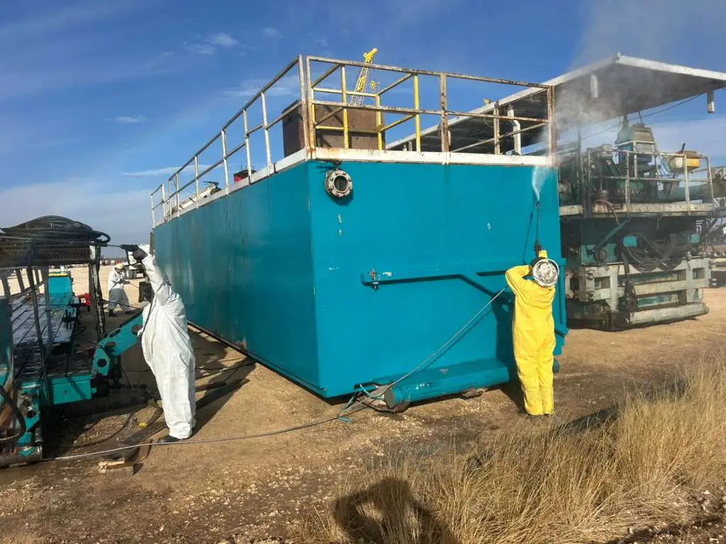 Two Gentex crew members in protective suits pressure washing a mud tank on location, spray coming off the wall of the tank