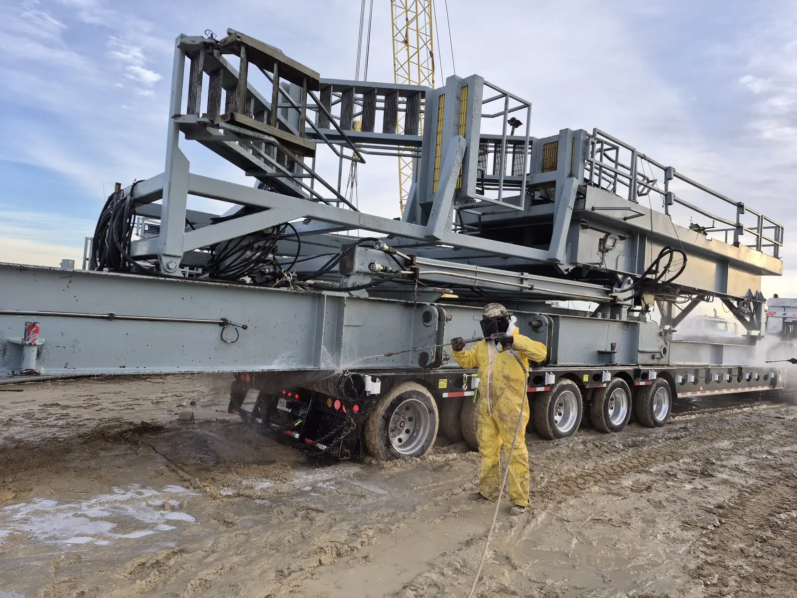 A Gentex operator in a chemical suit and face shield pressure washing a rig substructure loaded on a lowboy trailer