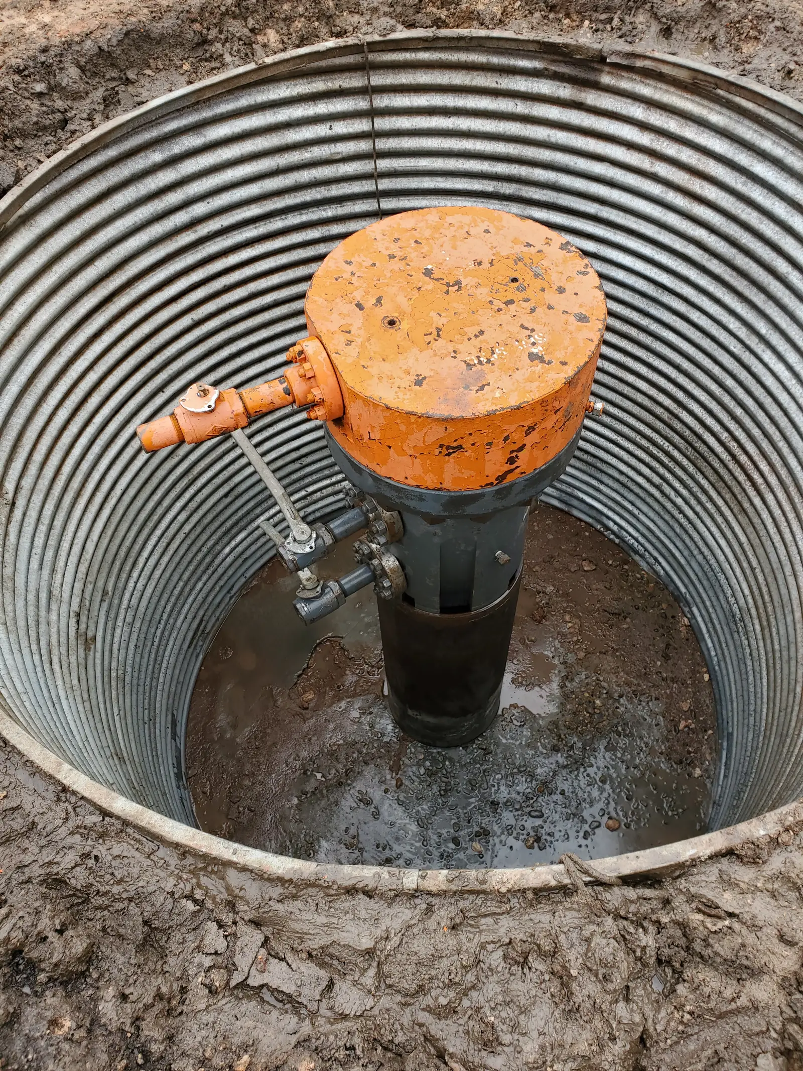 Wellhead cellar opened up for clean-out: corrugated cellar ring around an orange wellhead assembly with muddy water in the bottom